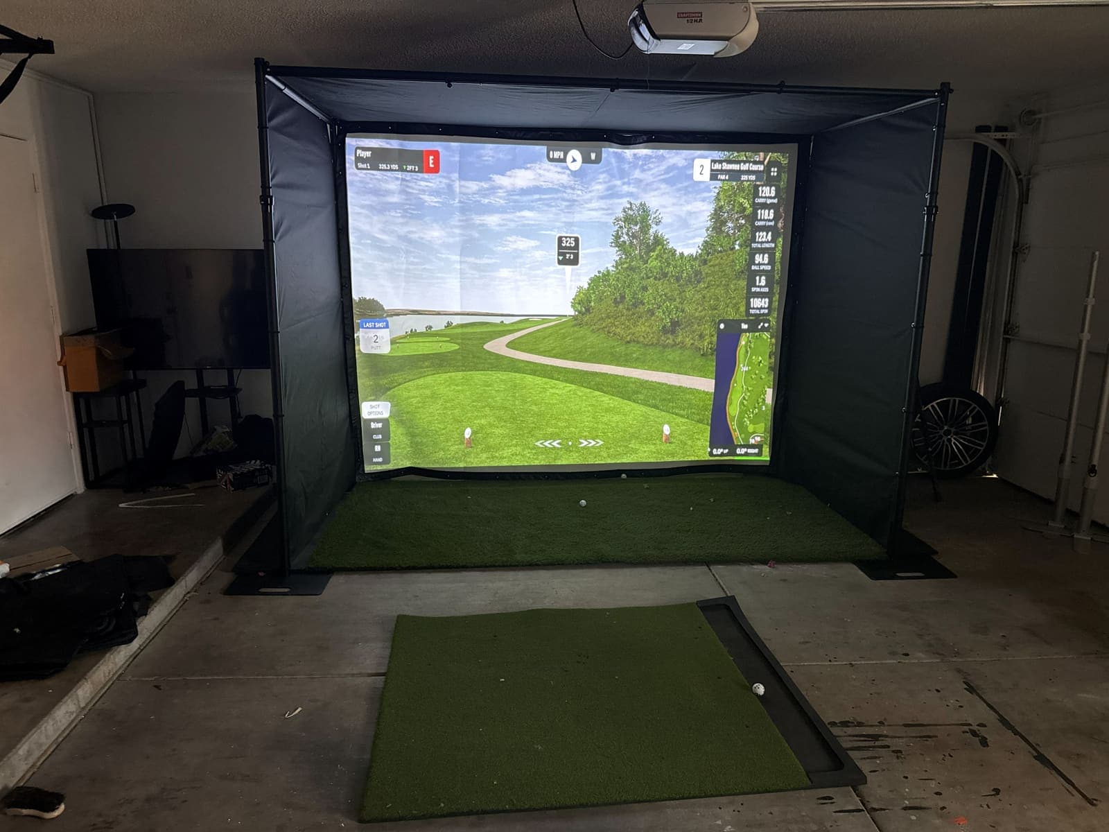 GSPro golf course projected on the impact screen inside the factory sample, a ball on the turf in front of it. Phone photo, October 11, 2026.